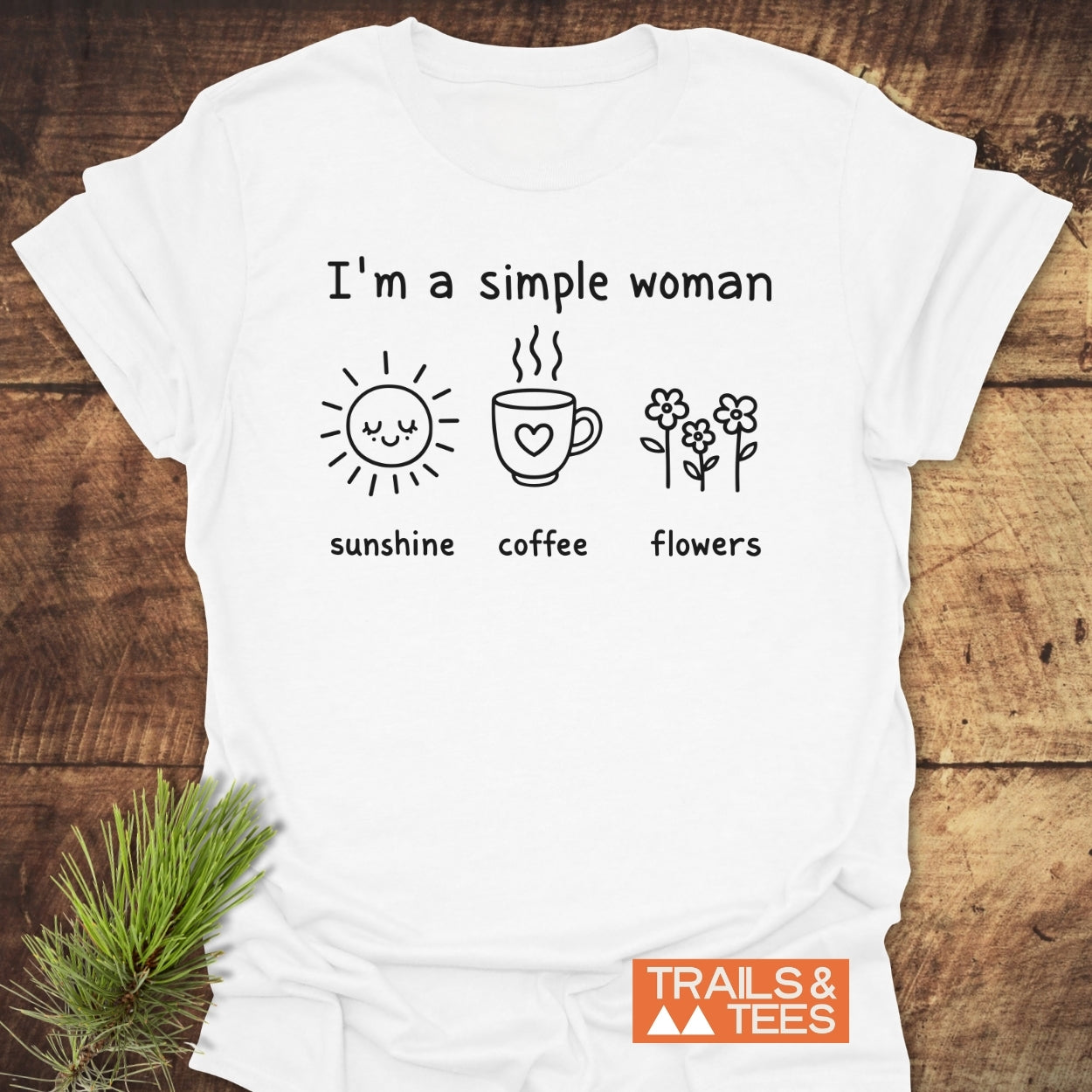 I'm A Simple Woman. Sunshine Coffee Flowers T-Shirt on wooden surface, featuring black icons of a sun, coffee mug, and flowers. 'TRAILS & TEES' logo in corner.