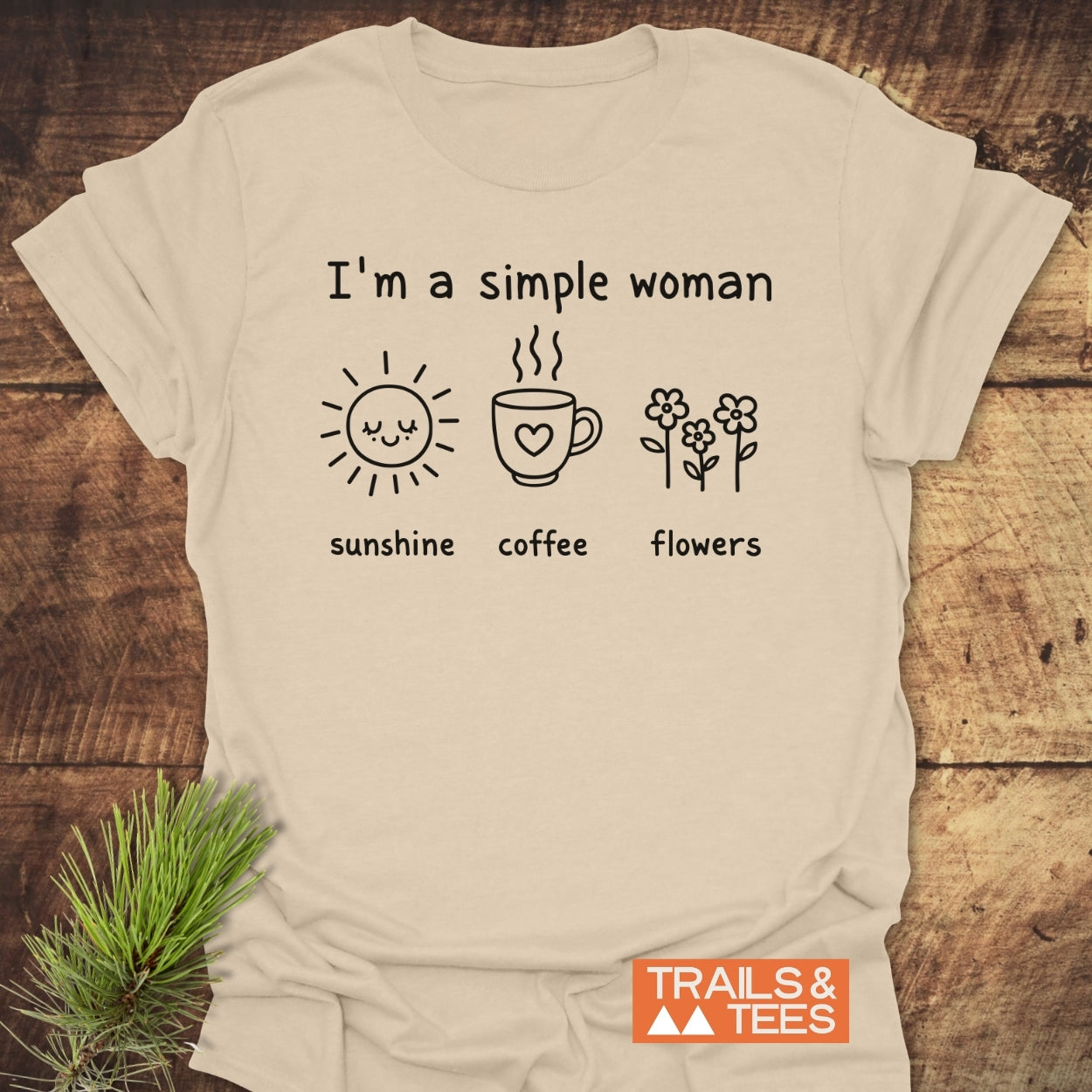 I'm A Simple Woman. Sunshine Coffee Flowers T-Shirt displayed on a wooden surface, featuring black line drawings of a sun, coffee mug, and flowers, with TRAILS & TEES logo in the corner.