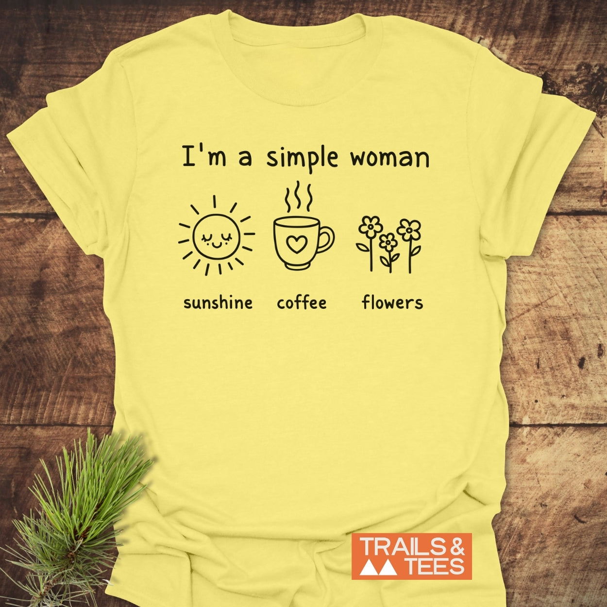 I'm A Simple Woman. Sunshine Coffee Flowers T-Shirt on wooden surface, features minimalist black line art with sun, coffee mug, flowers, and text. Includes small TRAILS & TEES logo in corner.