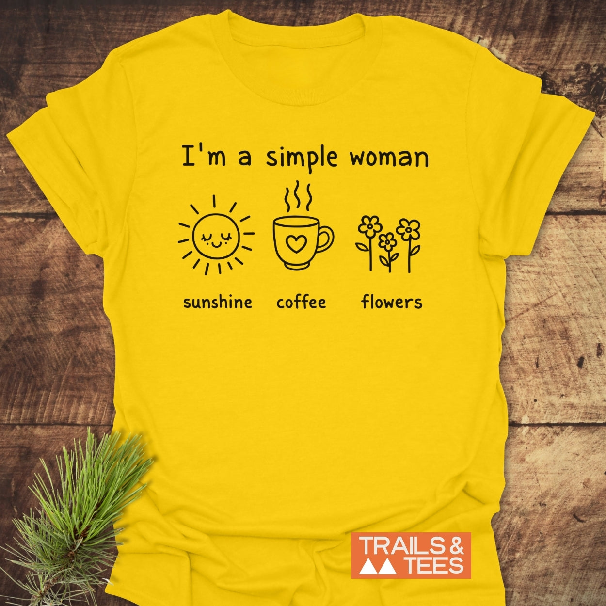 I'm A Simple Woman. Sunshine Coffee Flowers T-Shirt featuring a cheerful design with icons of a sun, coffee mug, and flowers. Laid on wood, includes TRAILS & TEES logo.
