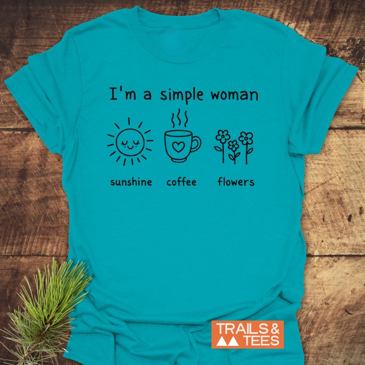 I'm A Simple Woman. Sunshine Coffee Flowers T-Shirt on wooden surface, featuring black print of sun, coffee, and flowers icons. Includes Trails & Tees logo, highlighting outdoor-inspired, durable design.