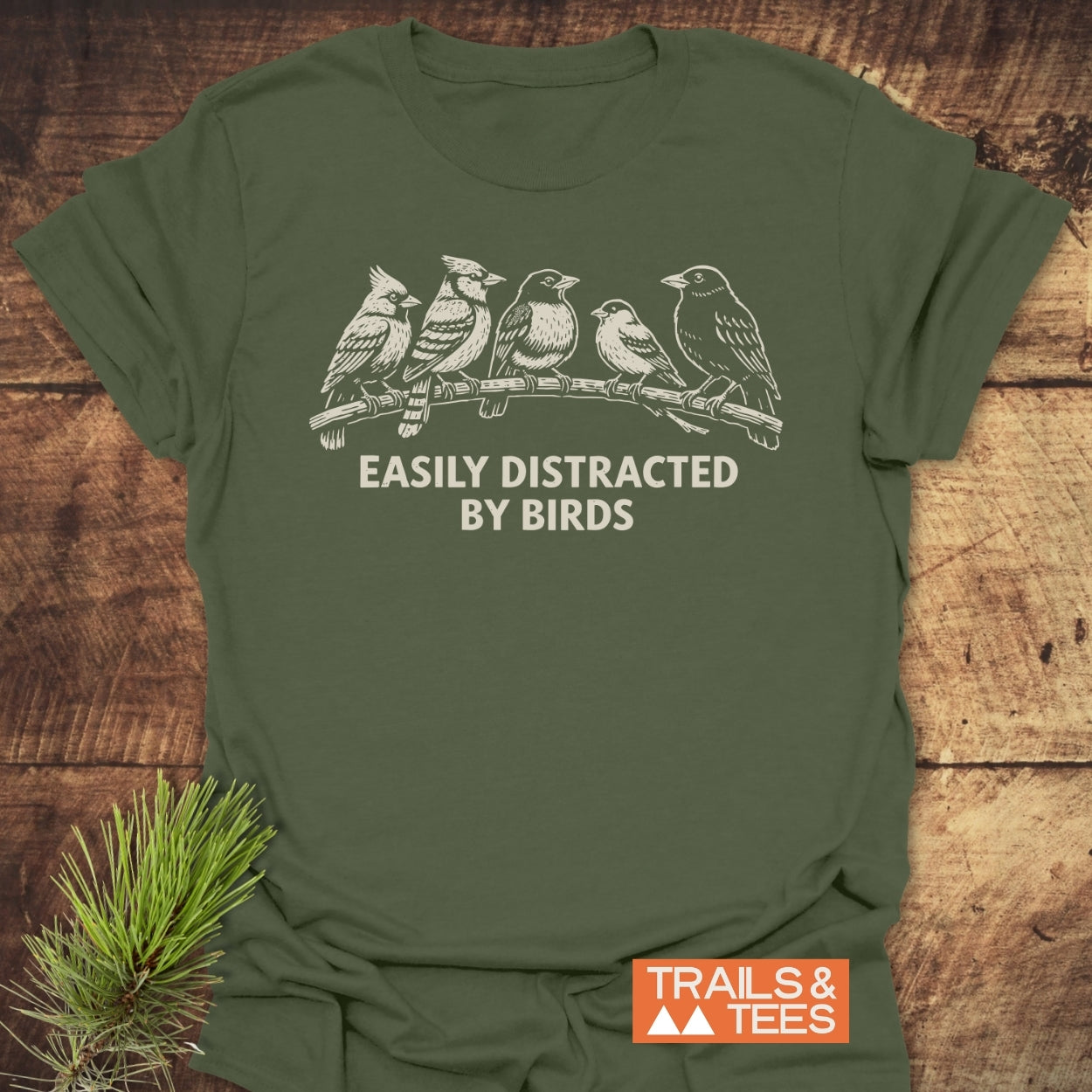 Easily Distracted By Birds T-Shirt featuring six birds on a branch and the phrase, laid on wood with pine sprig. Includes TRAILS & TEES logo, emphasizing outdoor adventure and comfort.