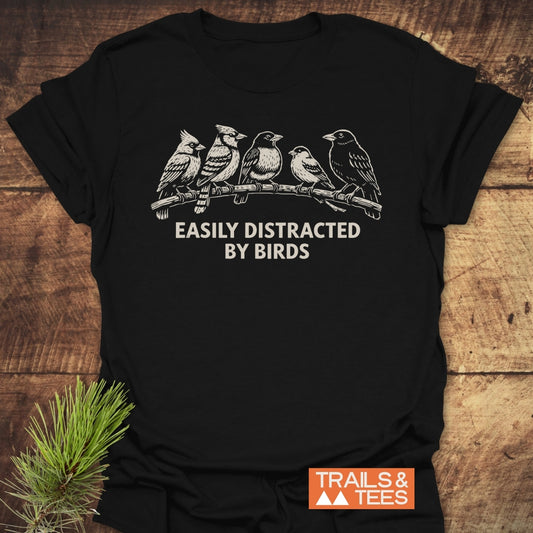 Easily Distracted By Birds T-Shirt on wood surface; features cream line drawing of birds on branch with bold slogan. Includes Trails & Tees logo, showcasing outdoor-inspired, comfortable design.