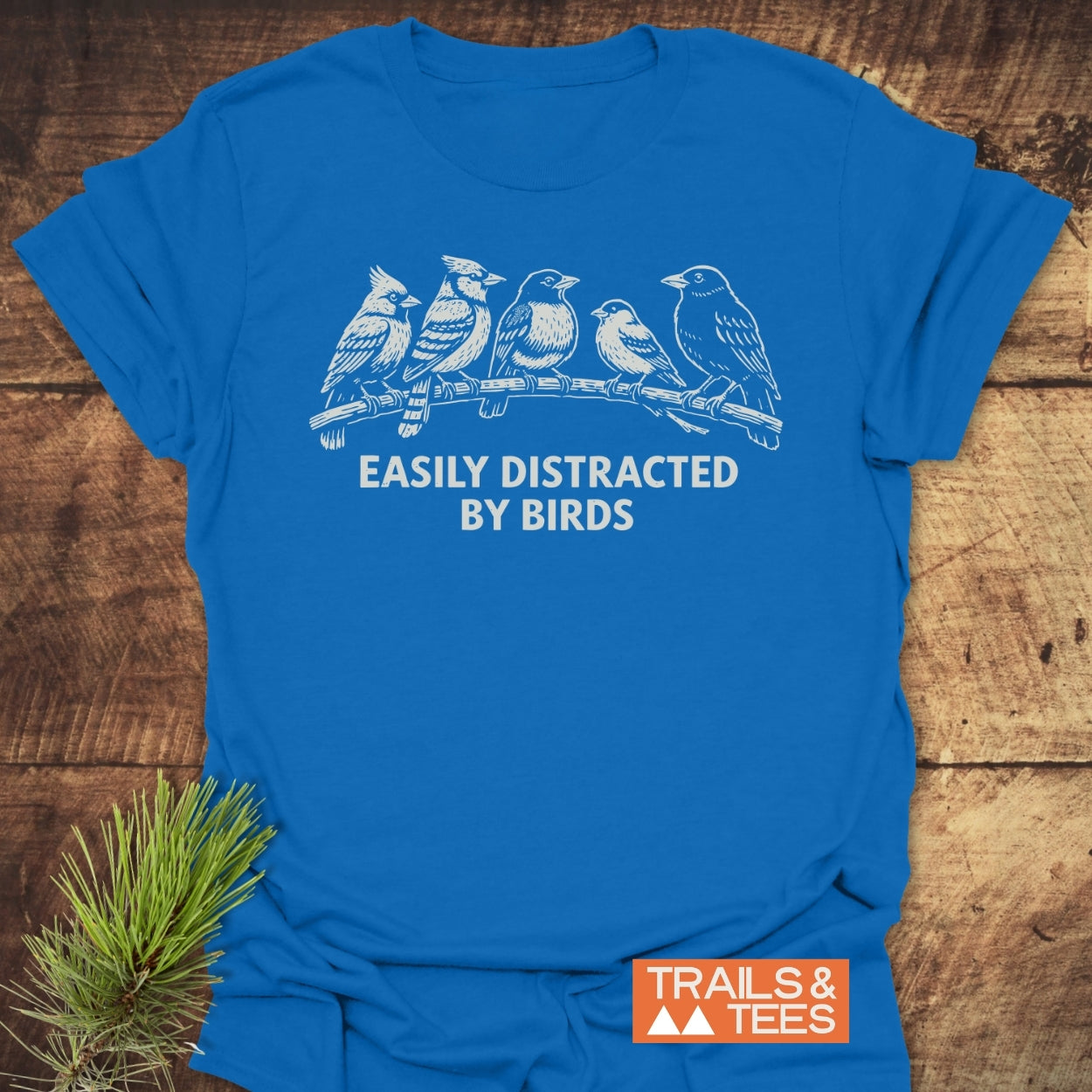 Easily Distracted By Birds T-Shirt featuring a white bird illustration and slogan on royal-blue cotton. Includes an orange TRAILS & TEES logo, styled with a pine sprig on wooden surface.