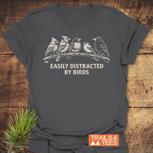 Easily Distracted By Birds T-Shirt featuring six songbirds illustration, slogan, and Trails & Tees logo on dark-gray fabric, styled with a pine sprig on a wooden surface.