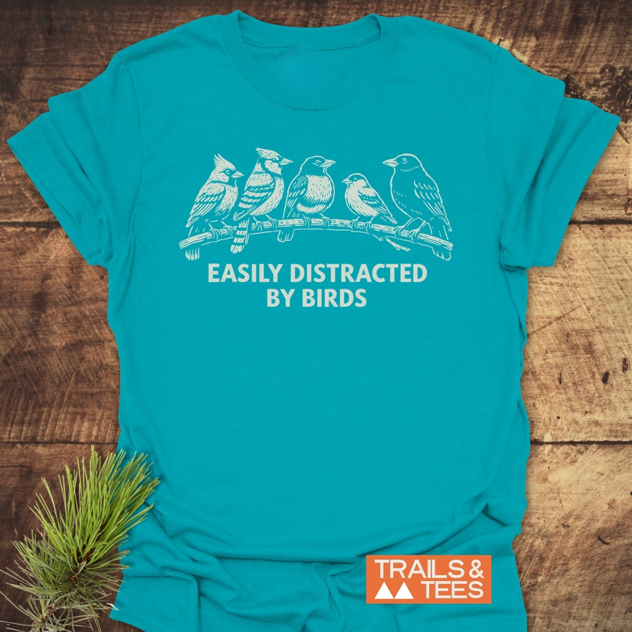 Easily Distracted By Birds T-Shirt featuring five white birds on a branch with slogan, laid on wood. Includes TRAILS & TEES logo and pine sprig, emphasizing outdoor adventure and comfort.