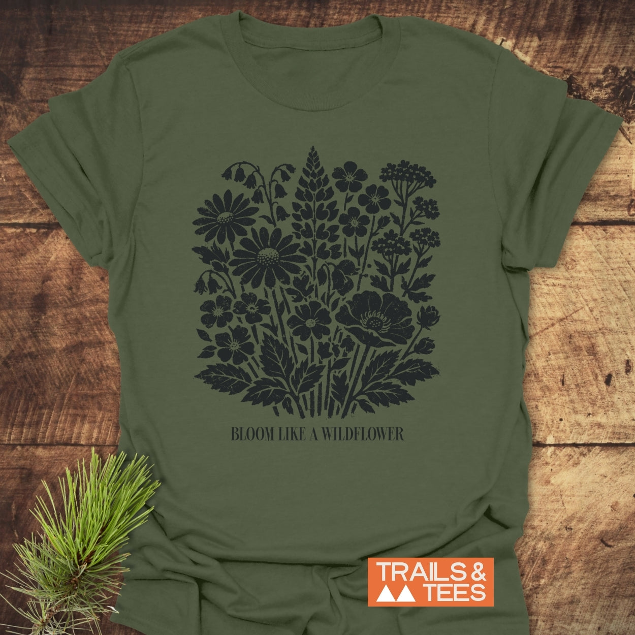 Bloom Like A Wildflower T-Shirt featuring a black wildflower graphic and inspiring text on an olive-green fabric. Styled with a pine sprig, showcasing its trail-ready, adventure-friendly design.