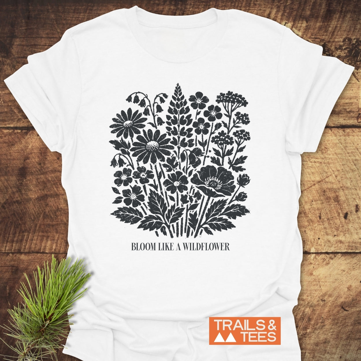 Bloom Like A Wildflower T-Shirt features a linocut-style wildflower design and slogan. Set on a wooden surface, it includes a “TRAILS & TEES” logo, highlighting outdoor adventure appeal.