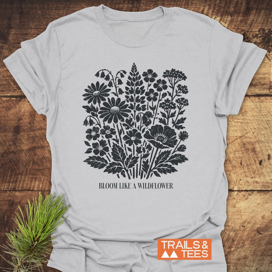 Bloom Like A Wildflower T-Shirt features a black-ink wildflower illustration with daisies, lupine, and poppies, plus the slogan beneath. Laid against rustic wood, highlighting its outdoor-inspired design.