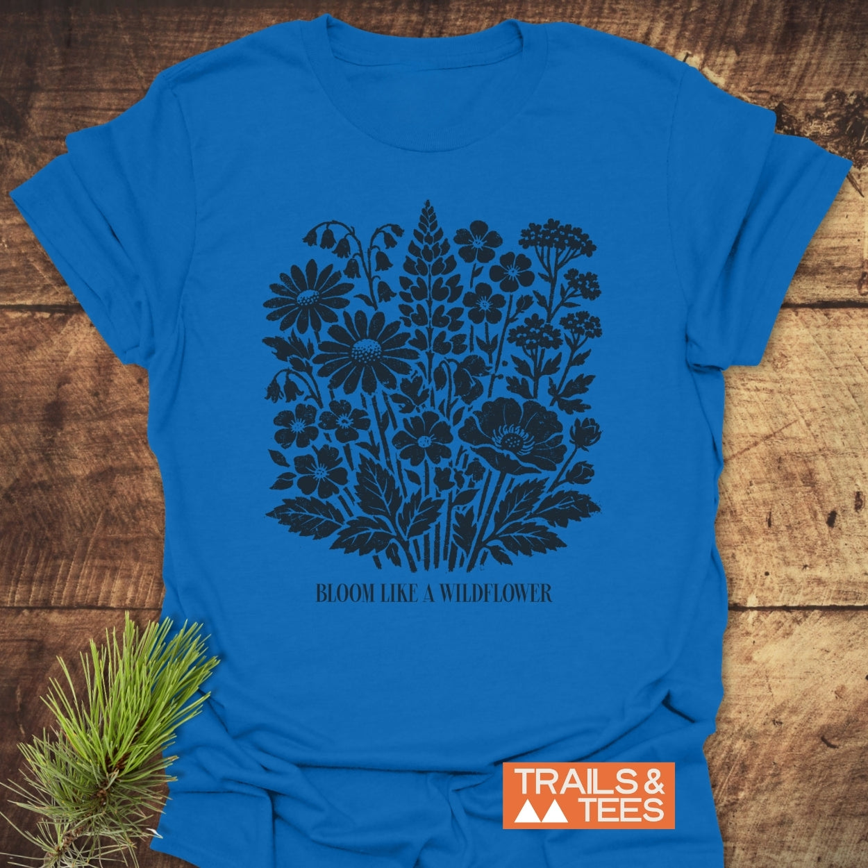 Bloom Like A Wildflower T-Shirt featuring a black wildflower silhouette and quote on a bright blue fabric, styled with a pine sprig, embodying outdoor adventure and comfort by Trails & Tees.