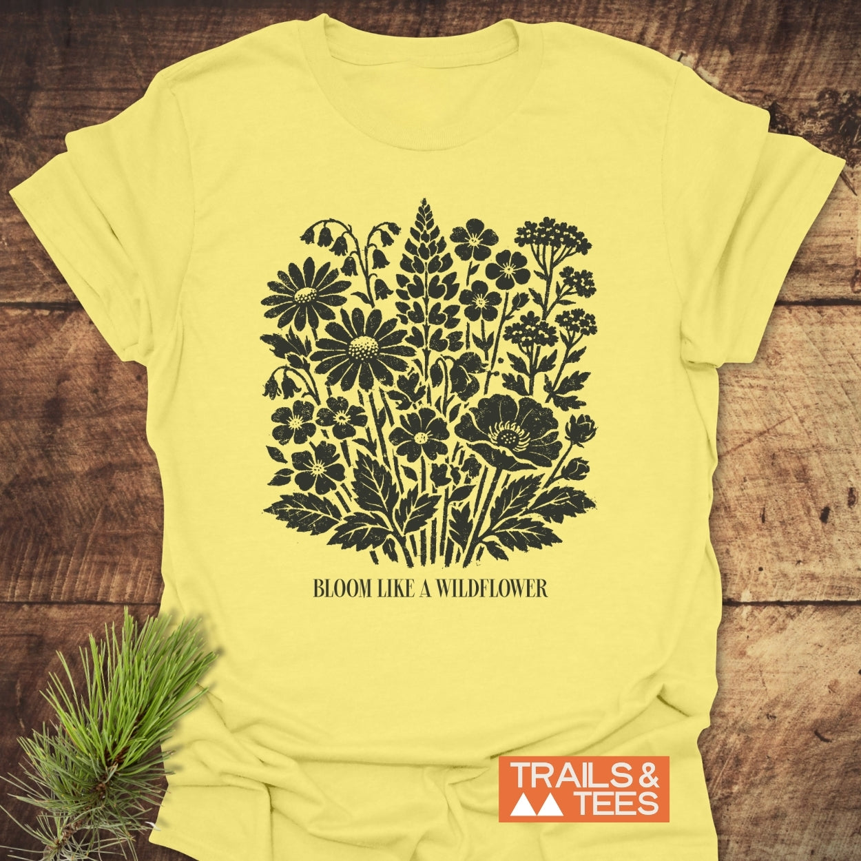 Bloom Like A Wildflower T-Shirt featuring a black vintage wildflower illustration and motto on pale yellow fabric, laid on rustic wood with an evergreen sprig; Trails & Tees logo visible.