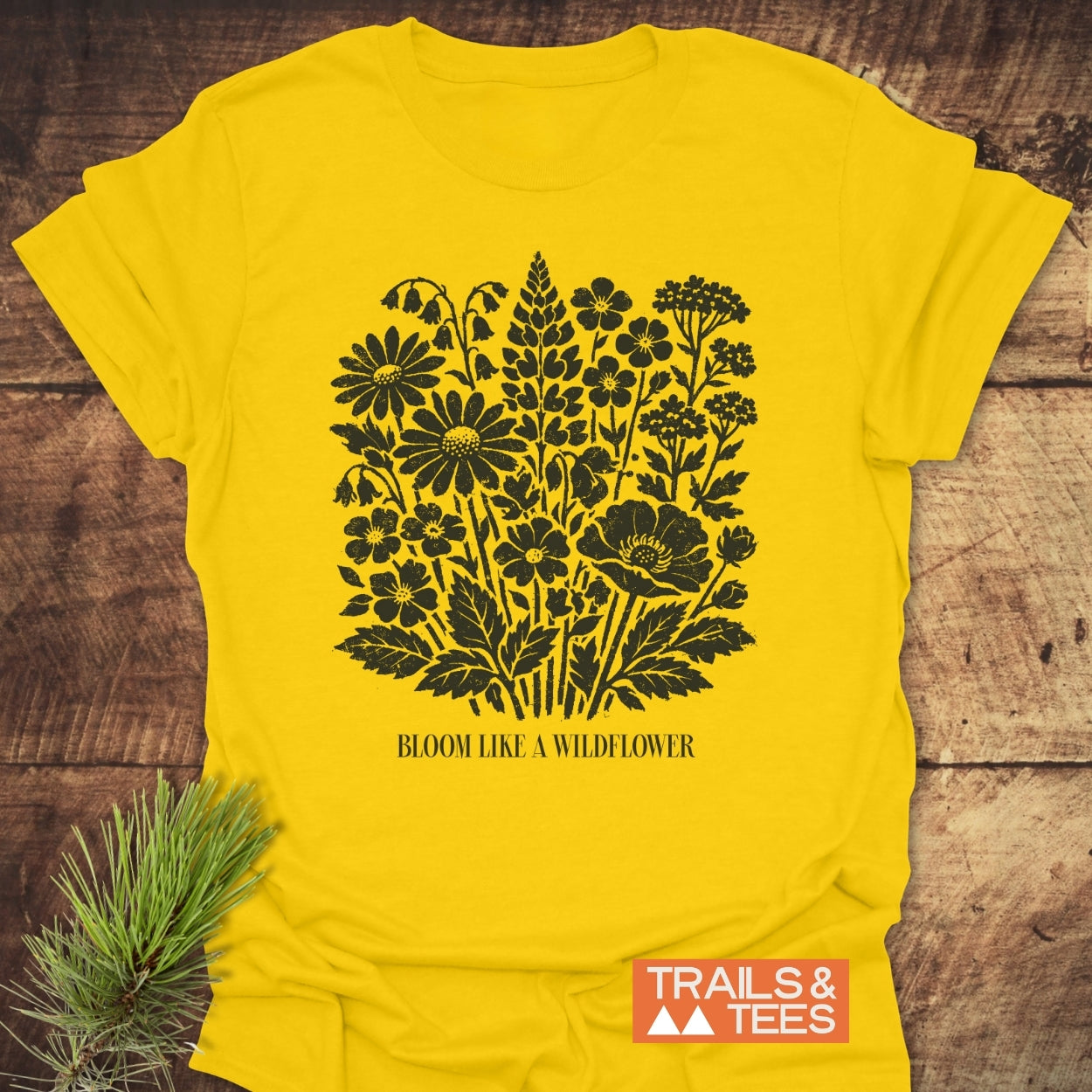 Bloom Like A Wildflower T-Shirt featuring black wildflower prints and slogan, laid on wood with pine sprig, embodies nature-inspired adventure. Crafted from soft ring-spun cotton by Trails & Tees.