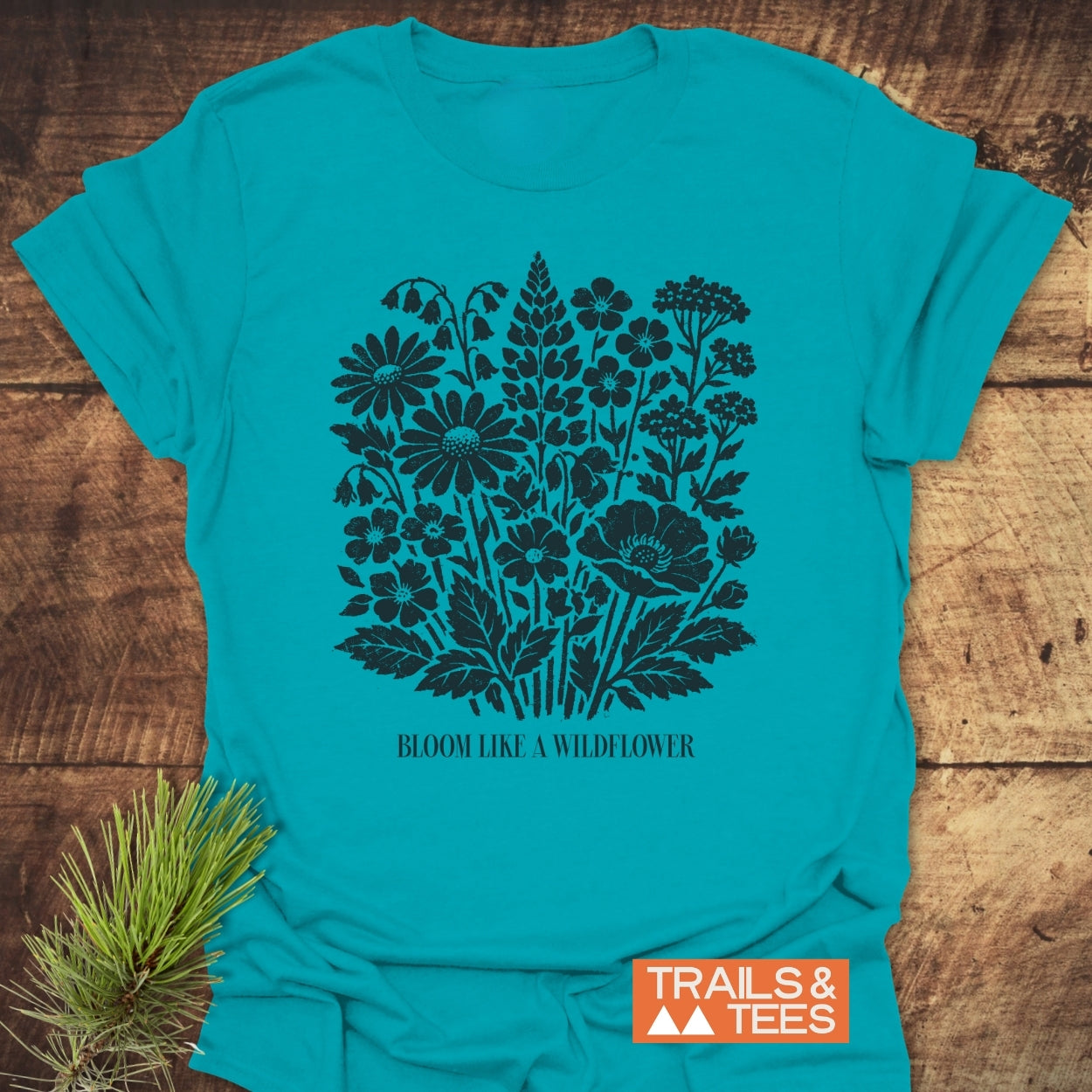 Bloom Like A Wildflower T-Shirt featuring a stylized wildflower silhouette and slogan, crafted from soft, lightweight ring-spun cotton. Includes TRAILS & TEES logo, ideal for outdoor adventure lovers.