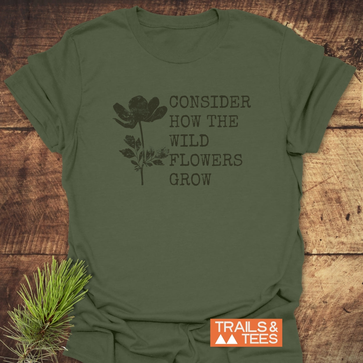 Consider How The Wild Flowers Grow T-Shirt on rustic wood, featuring a black wildflower silhouette and phrase, with Trails & Tees badge, embodying outdoor adventure spirit. Pine sprig accentuates nature theme.