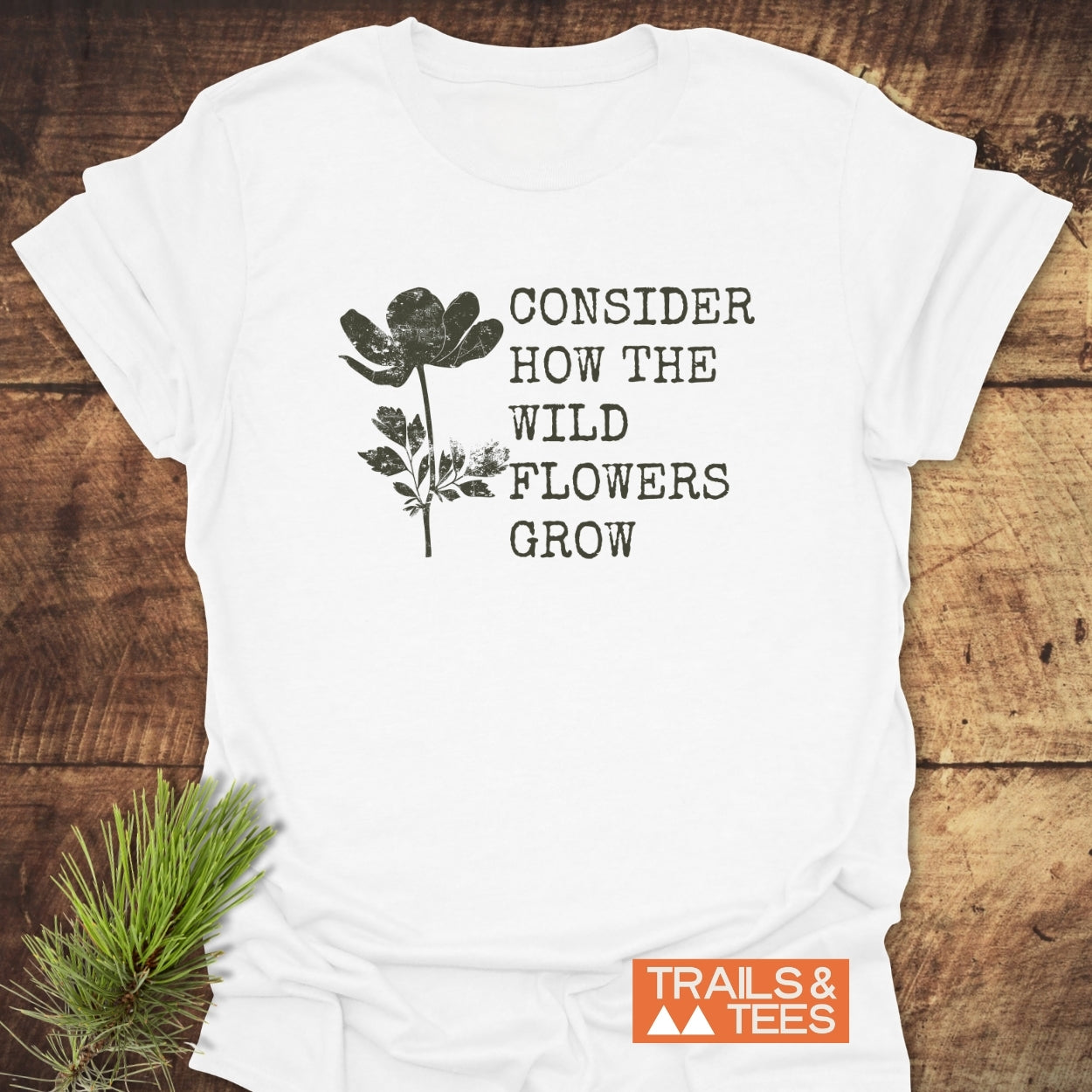 Consider How The Wild Flowers Grow T-Shirt on rustic wood, featuring a distressed wildflower silhouette and quote. Includes a small orange TRAILS & TEES logo, emphasizing outdoor-inspired design.