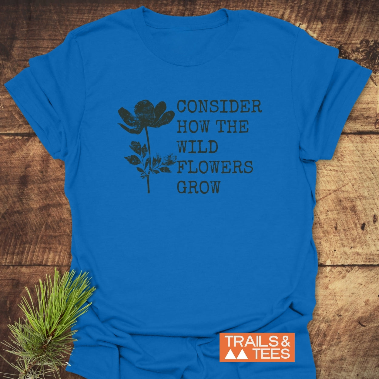 Consider How The Wild Flowers Grow T-Shirt featuring a black wildflower line drawing with text, laid on wood. Includes “TRAILS & TEES” logo, highlighting its outdoor, adventure-ready design.