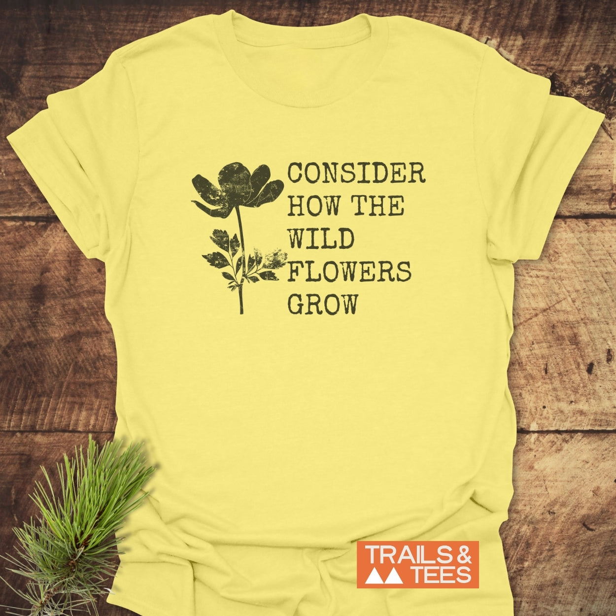 Consider How The Wild Flowers Grow T-Shirt, featuring a black wildflower graphic and phrase, laid on wood. Includes a small “TRAILS & TEES” logo. Pine sprig accents the scene.