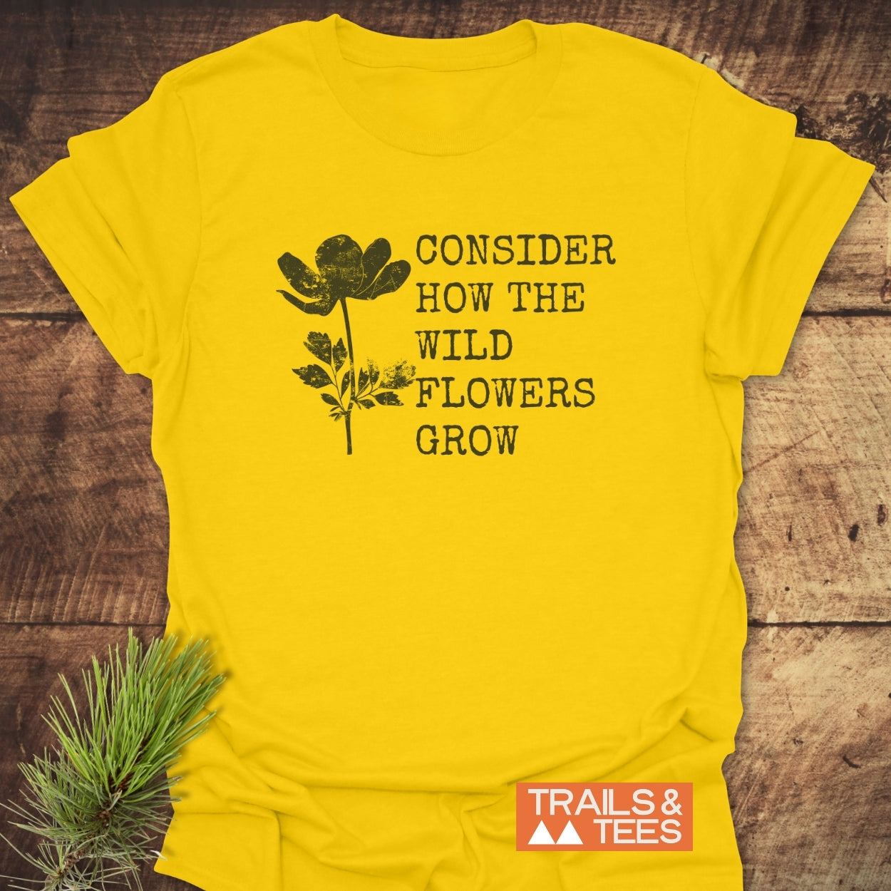Bright yellow Consider How The Wild Flowers Grow T-Shirt with black wildflower drawing and rustic text. Styled on wood with evergreen sprig. Features Trails & Tees logo, emphasizing outdoor-inspired, durable comfort.