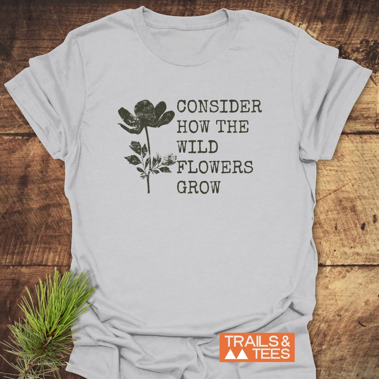 Consider How The Wild Flowers Grow T-Shirt displayed on wood, featuring a distressed wildflower illustration and the phrase. Includes “TRAILS & TEES” logo, emphasizing comfort and durability for outdoor enthusiasts.