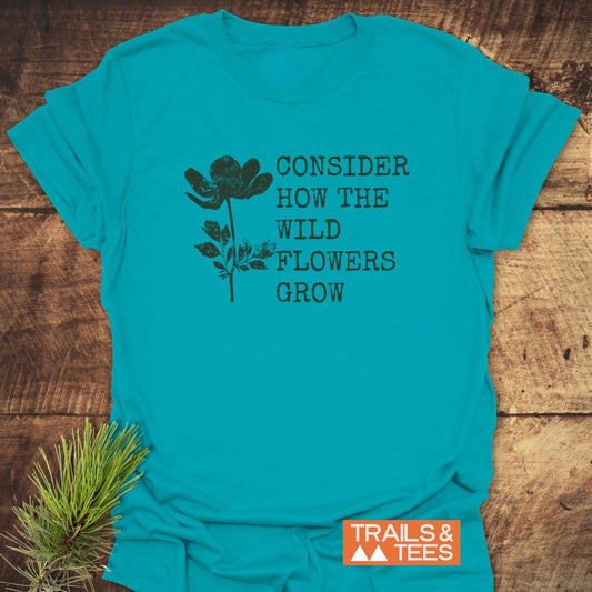 Consider How The Wild Flowers Grow T-Shirt features a black wildflower silhouette and quote on a turquoise-teal background, complemented by Trails & Tees logo. Pine needles accentuate the rustic presentation.