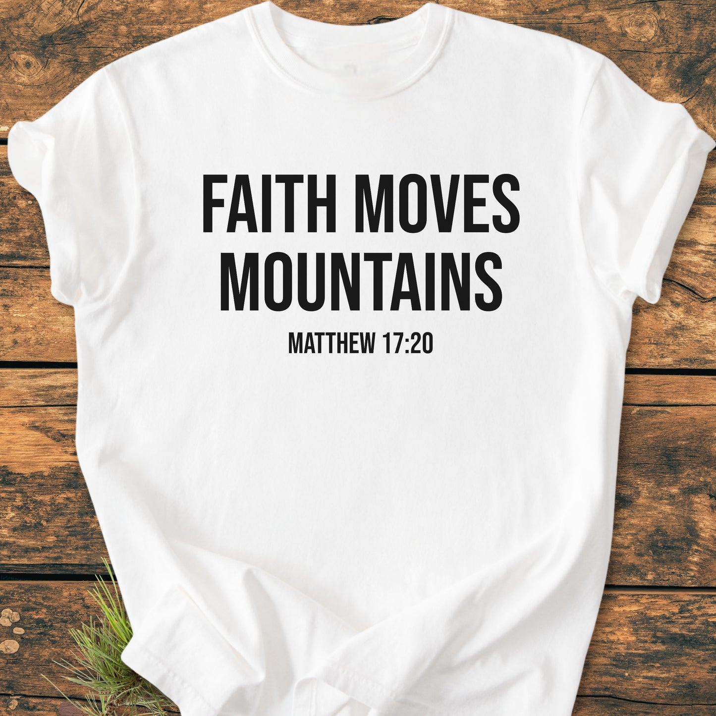 Faith Moves Mountains - Vintage Wash Tee, featuring bold black text on a white T-shirt, showcases FAITH MOVES MOUNTAINS and MATTHEW 17:20, laid out on a wooden surface.