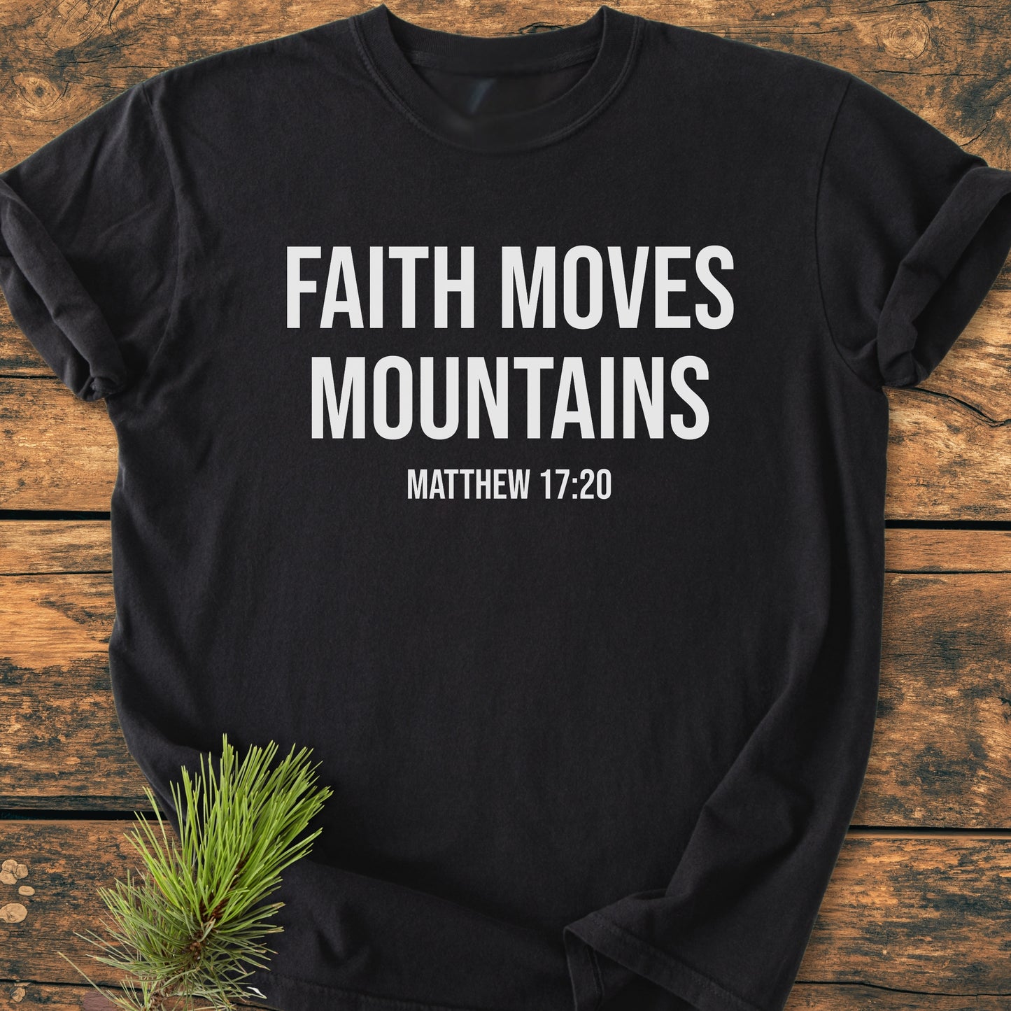 Faith Moves Mountains - Vintage Wash Tee featuring bold white text on a black, short-sleeved, soft-washed cotton shirt, laid on wood with a pine sprig accentuating its rustic presentation.