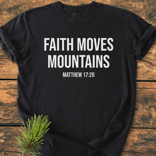 Faith Moves Mountains - Vintage Wash Tee featuring bold white text on a black, short-sleeved, soft-washed cotton shirt, laid on wood with a pine sprig accentuating its rustic presentation.