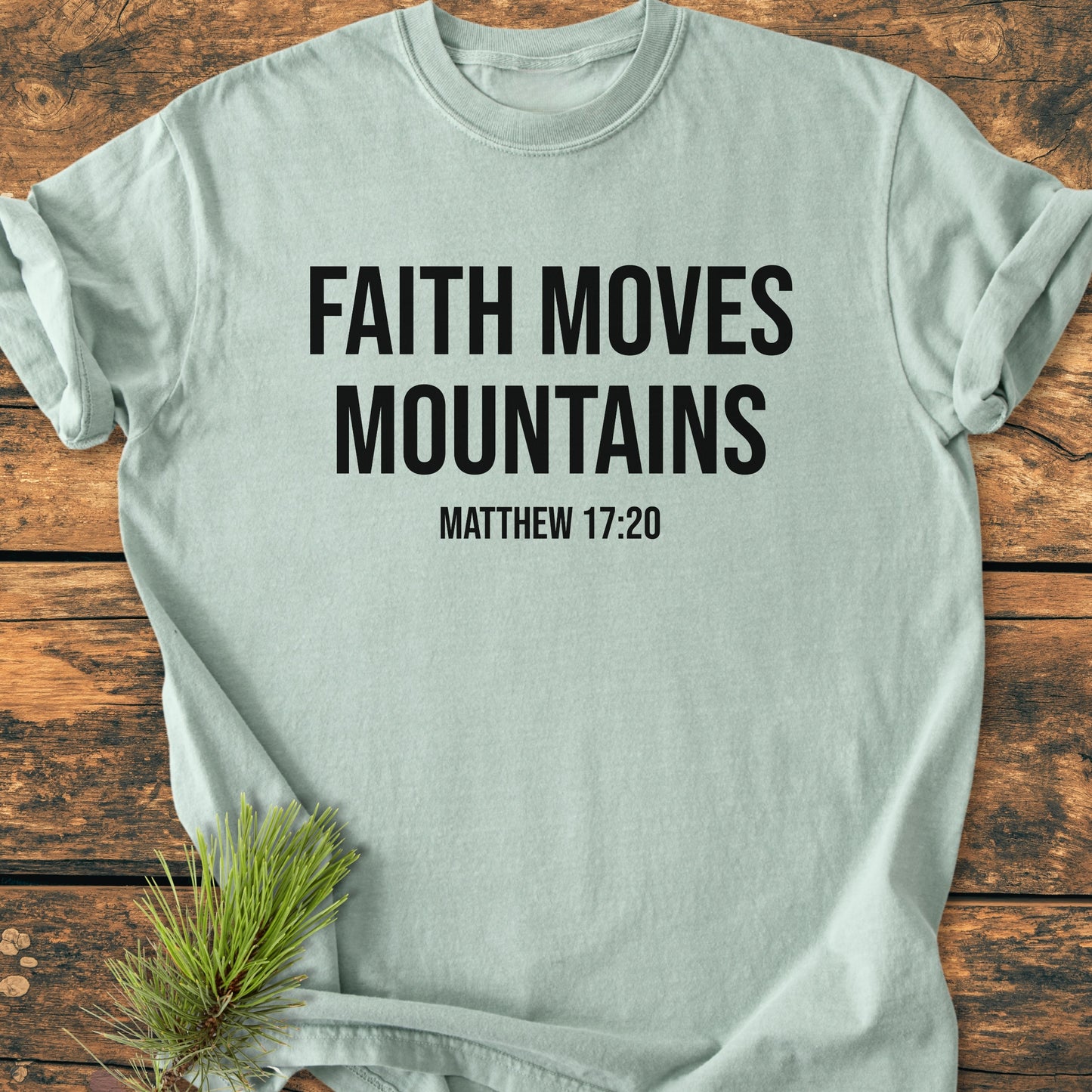 Faith Moves Mountains - Vintage Wash Tee featuring bold slogan and scripture on soft fabric, laid on wood. Pine sprig accentuates this relaxed fit, pre-shrunk, 100% ring-spun cotton T-shirt.