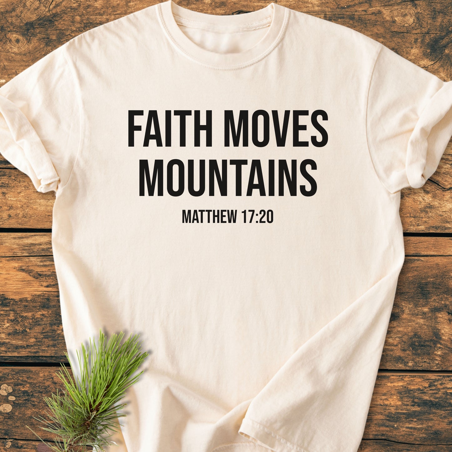 Faith Moves Mountains - Vintage Wash Tee displayed on a wooden surface, features bold black lettering and a biblical reference. Made of soft, durable, garment-dyed 100% ring-spun cotton for a relaxed fit.