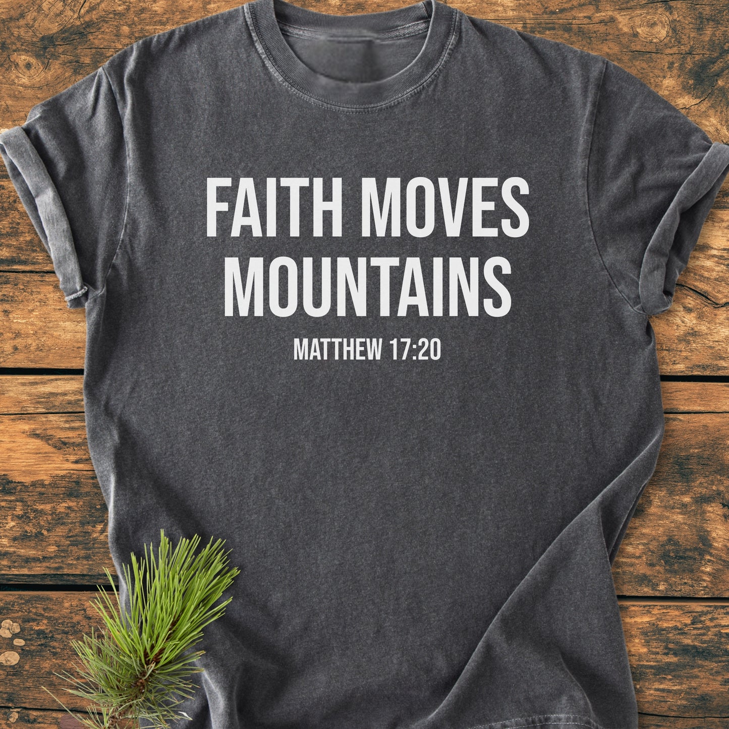 Faith Moves Mountains - Vintage Wash Tee: A dark gray T-shirt with FAITH MOVES MOUNTAINS and MATTHEW 17:20 printed in white, featuring rolled sleeves, laid on a wooden surface with pine needles.