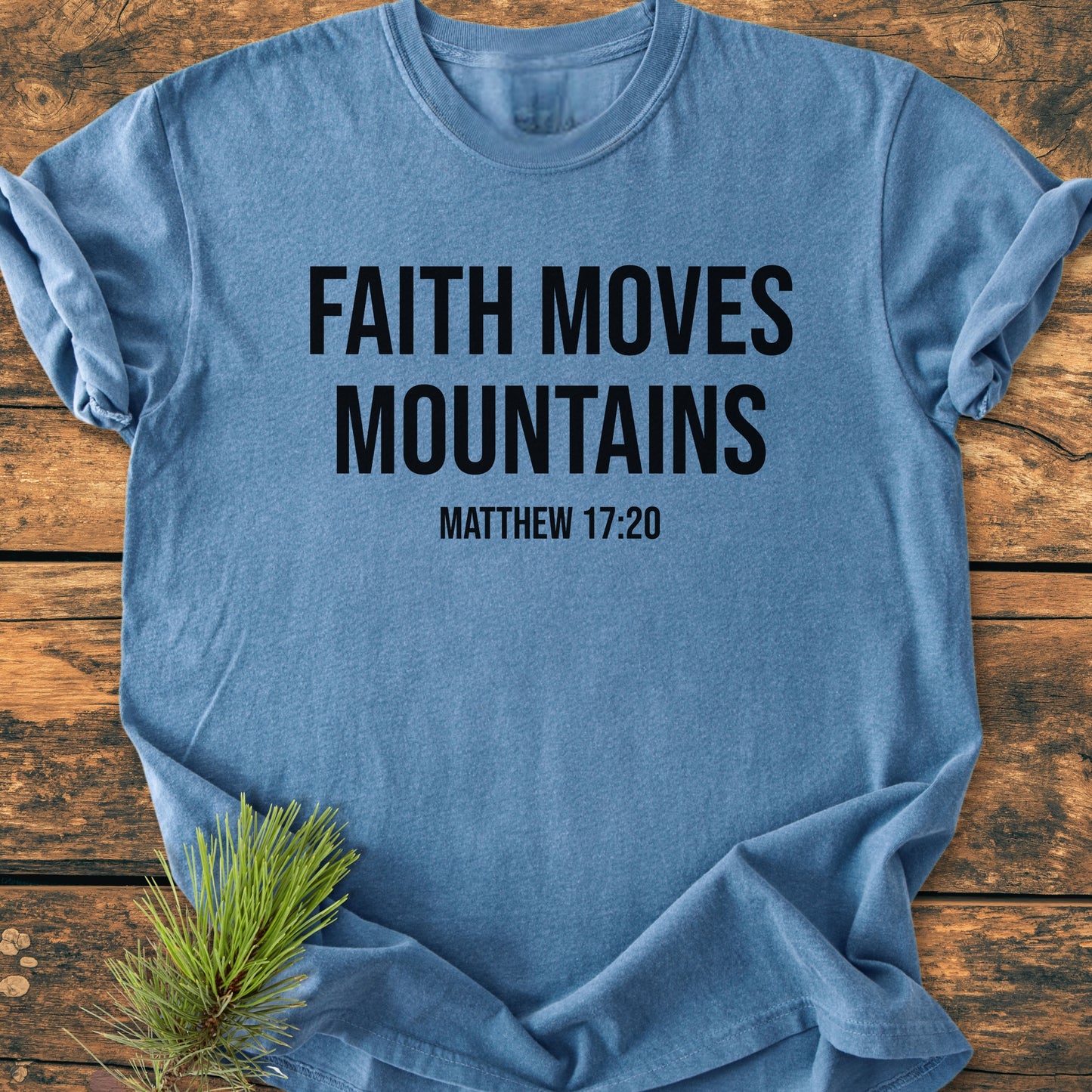 Faith Moves Mountains - Vintage Wash Tee displayed flat on wood, features bold black text and Matthew 17:20. Soft-blue, garment-dyed, 100% cotton, emphasizing comfort and durability. Pine needles accent the scene.