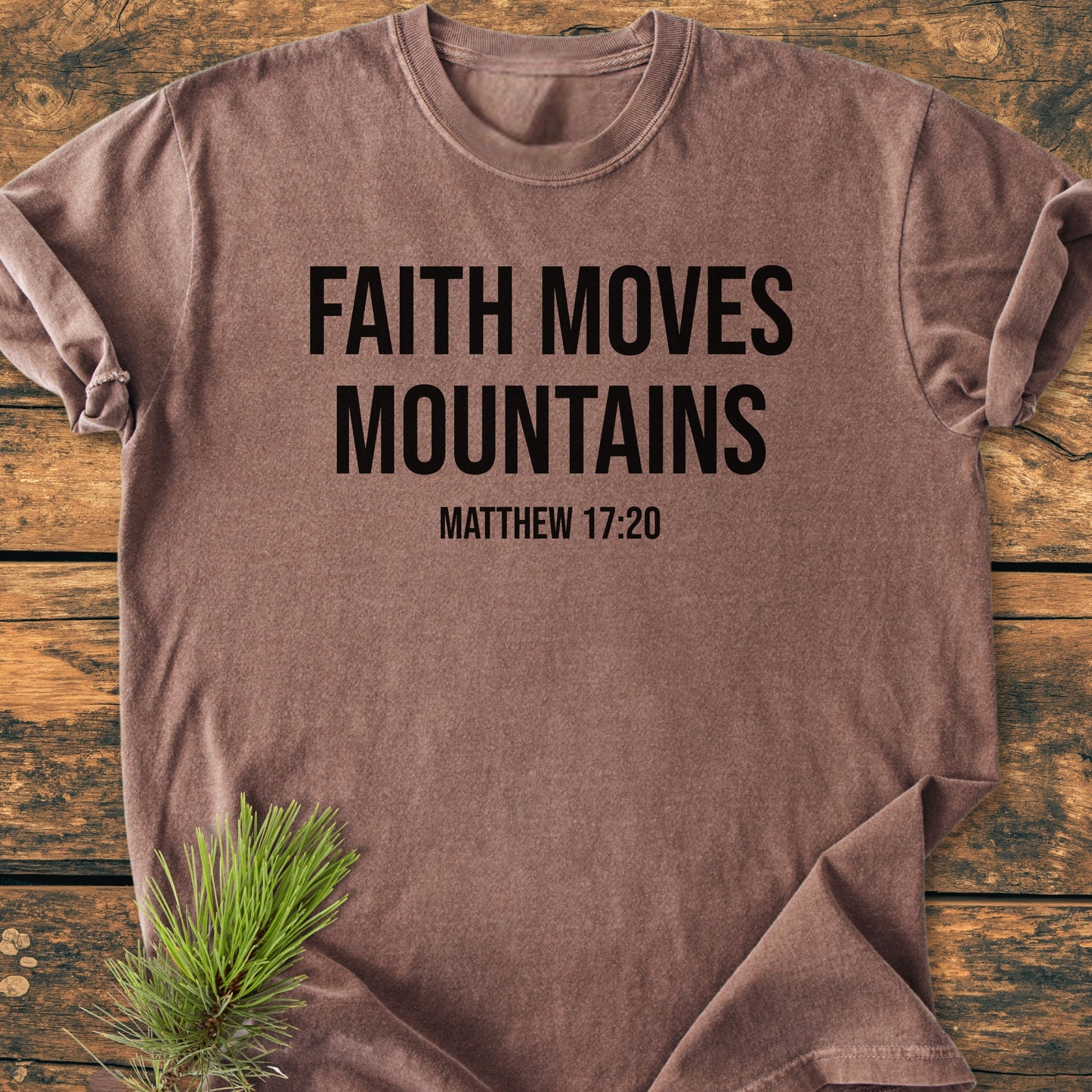 Faith Moves Mountains - Vintage Wash Tee features bold black text and scripture on a brown garment-dyed T-shirt. Laid flat on a wooden surface with a pine branch accent.