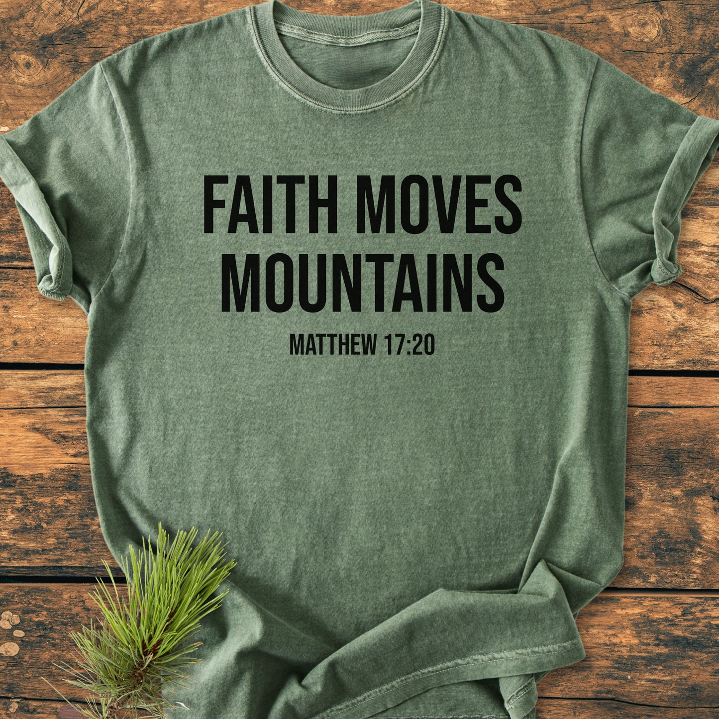 Heather-green Faith Moves Mountains - Vintage Wash Tee on wood surface, features bold black lettering and scripture reference. Pine sprig beside shirt enhances rustic appeal.