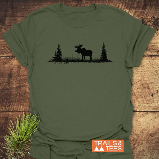 Lone Moose T-Shirt flat-lay: olive-green with a black moose silhouette between pine trees, laid on rustic wood. Features TRAILS & TEES logo. Crafted from soft, lightweight ring-spun cotton.
