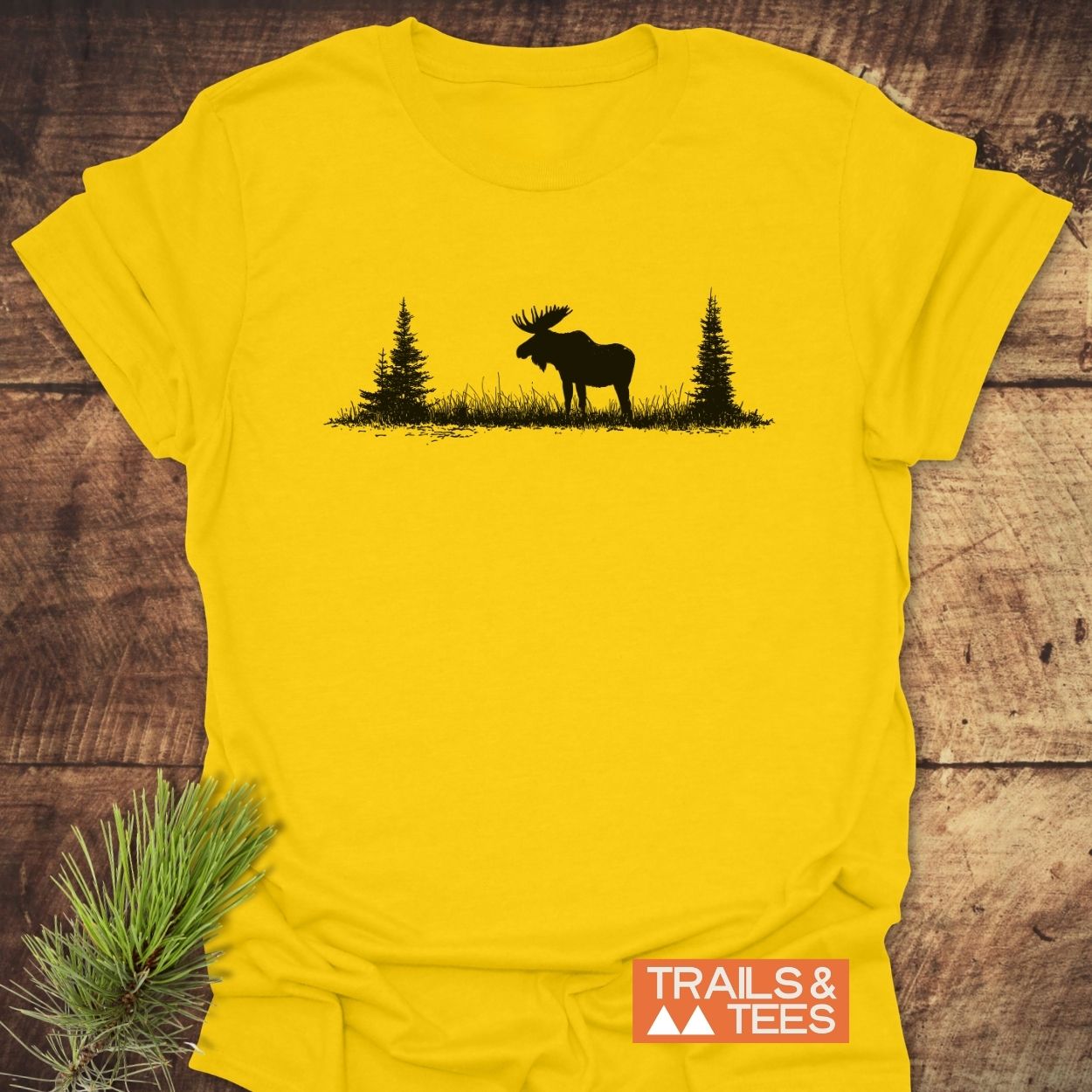 Lone Moose T-Shirt features a black moose silhouette with trees on a bright yellow shirt, laid flat. Includes a TRAILS & TEES logo, embodying an outdoorsy theme with soft, durable fabric.