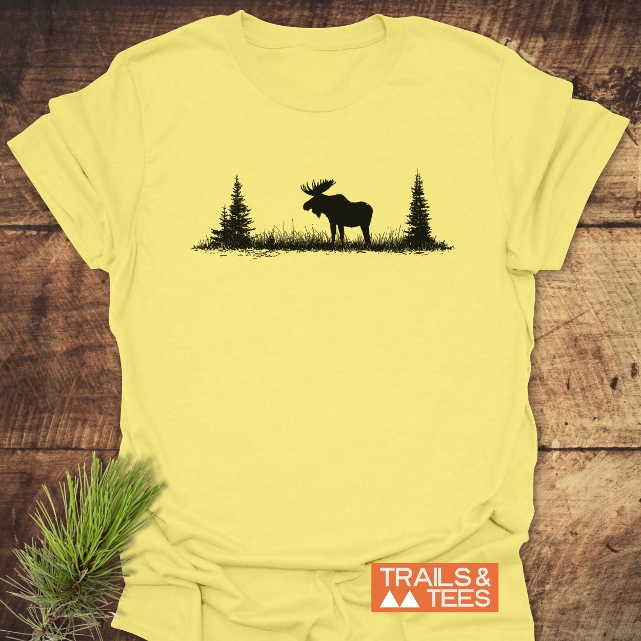 Lone Moose T-Shirt features a black moose silhouette with pine trees on a pale yellow fabric, highlighting outdoor adventure. Includes a TRAILS & TEES logo and pine sprig accent.