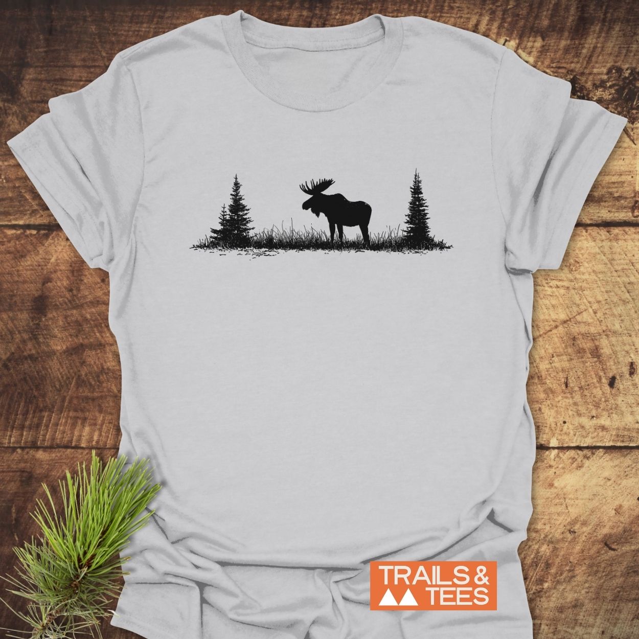 Lone Moose T-Shirt: Light-gray tee featuring a black moose silhouette with pine trees, crafted from soft cotton for comfort. Includes an orange TRAILS & TEES label and a pine branch accent.