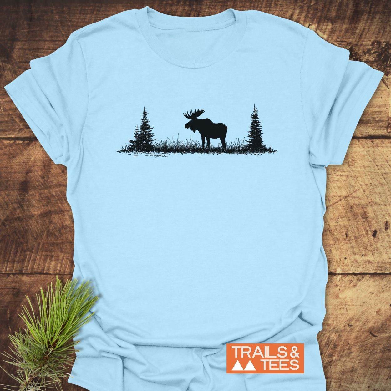 Lone Moose T-Shirt featuring a nature silhouette of a moose and pine trees, laid flat on wood with brand logo “TRAILS & TEES,” styled with a pine sprig for an outdoor feel.