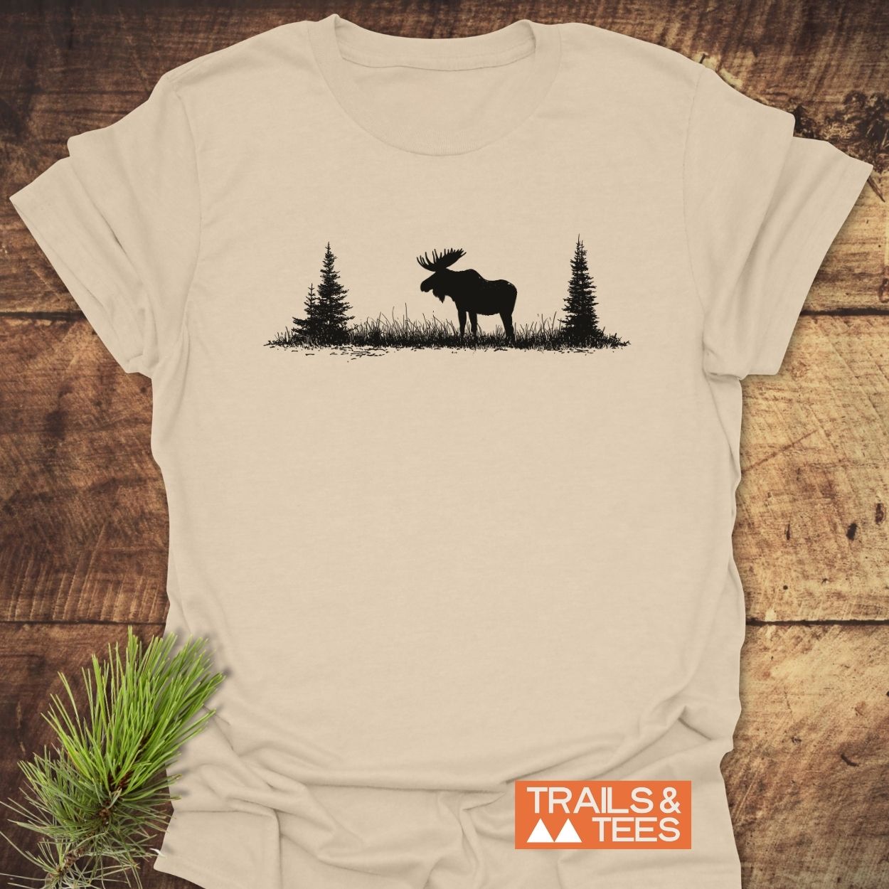 Lone Moose T-Shirt featuring a moose silhouette with pine trees on light-beige cotton, styled with a pine sprig. Ideal for outdoor enthusiasts seeking comfort and adventure.