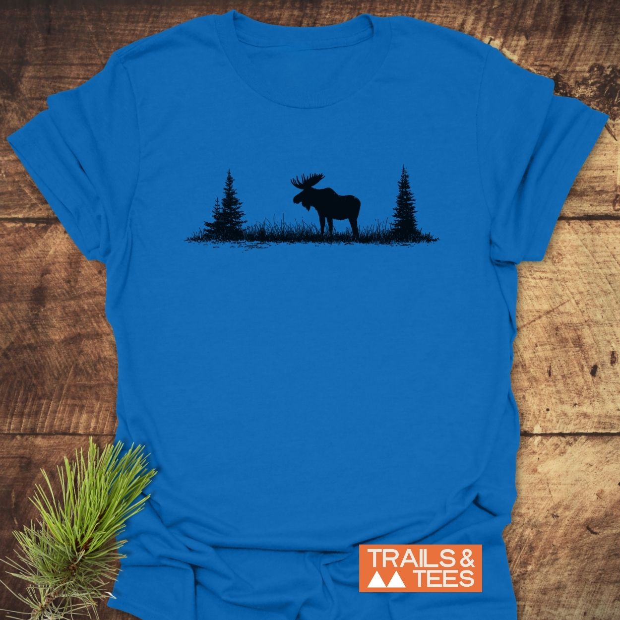 Lone Moose T-Shirt featuring a moose and evergreen trees silhouette, with an orange Trails & Tees logo, laid out with a pine sprig, highlighting its outdoor-inspired design.