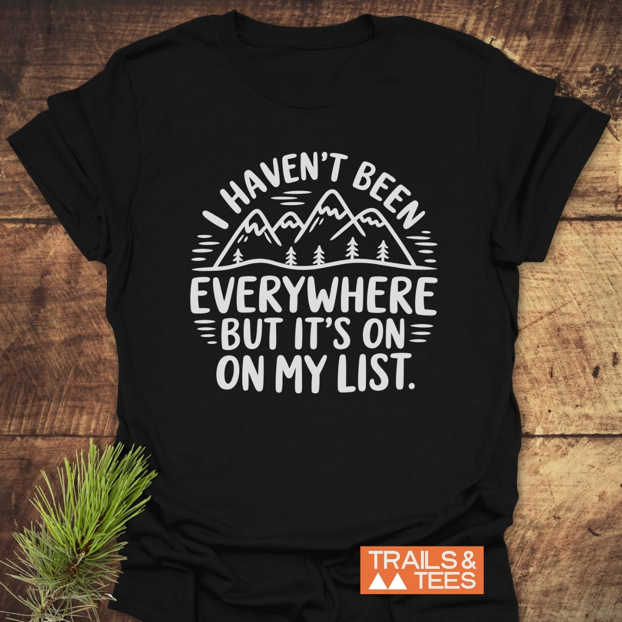On My List T-Shirt featuring a white mountain-and-trees graphic and slogan, laid flat with a pine sprig beside it, and TRAILS & TEES logo in the corner, highlighting outdoor adventure spirit.