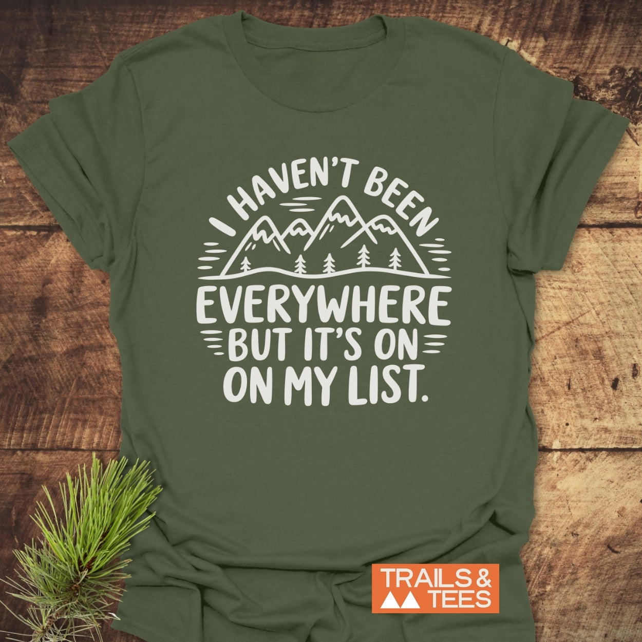 Olive-green On My List T-Shirt featuring a mountain-and-pine tree graphic and adventurous quote, styled with a pine sprig. Includes Trails & Tees logo, emphasizing outdoor-inspired comfort and durability.