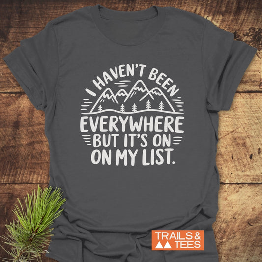 On My List T-Shirt features a white line-drawing of mountains and trees with an adventurous quote, set on soft charcoal-gray fabric, ideal for outdoor enthusiasts.