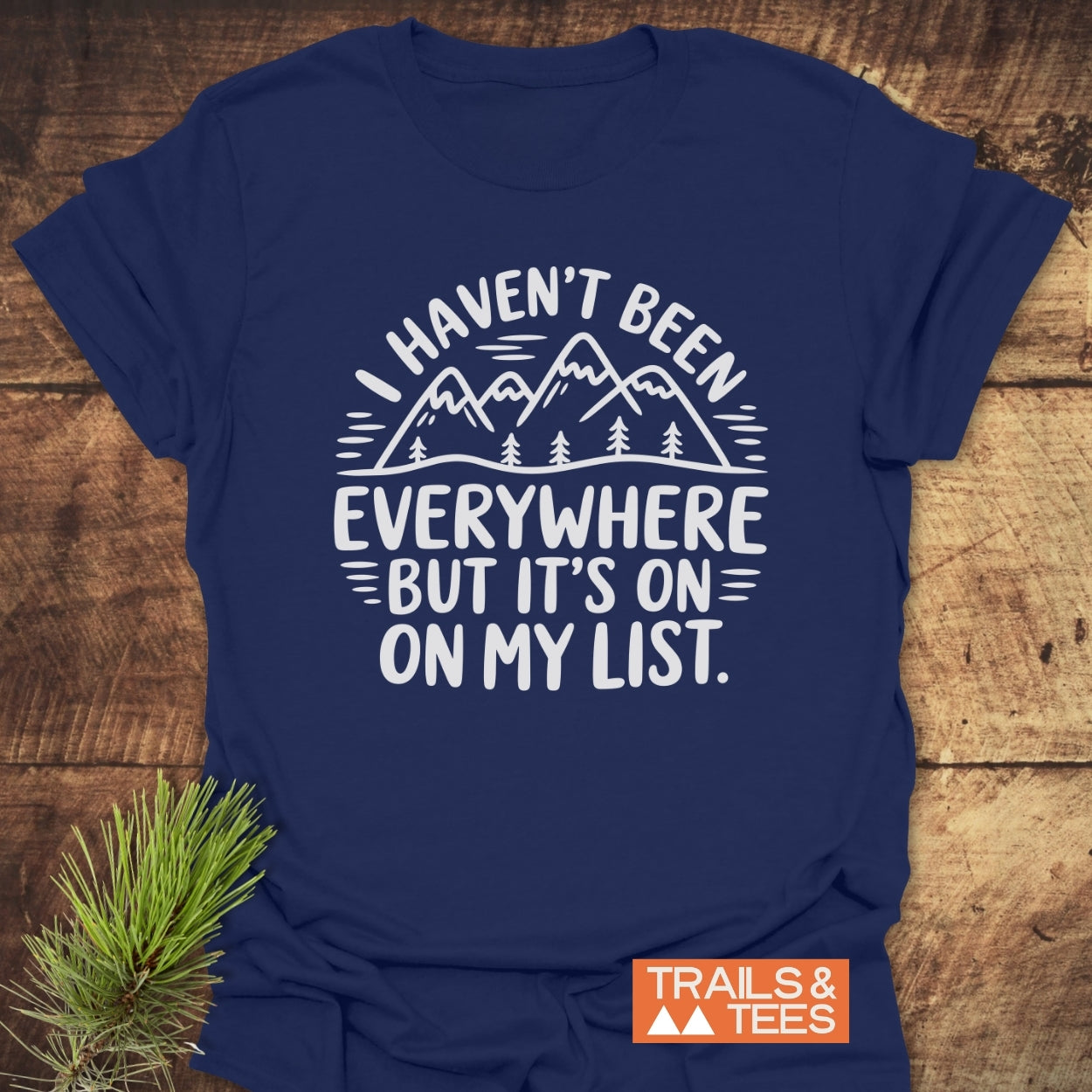 Navy-blue On My List T-Shirt featuring white mountain graphic and slogan, laid flat with pine sprig. Includes orange and white “TRAILS & TEES” logo, highlighting adventure and comfort.