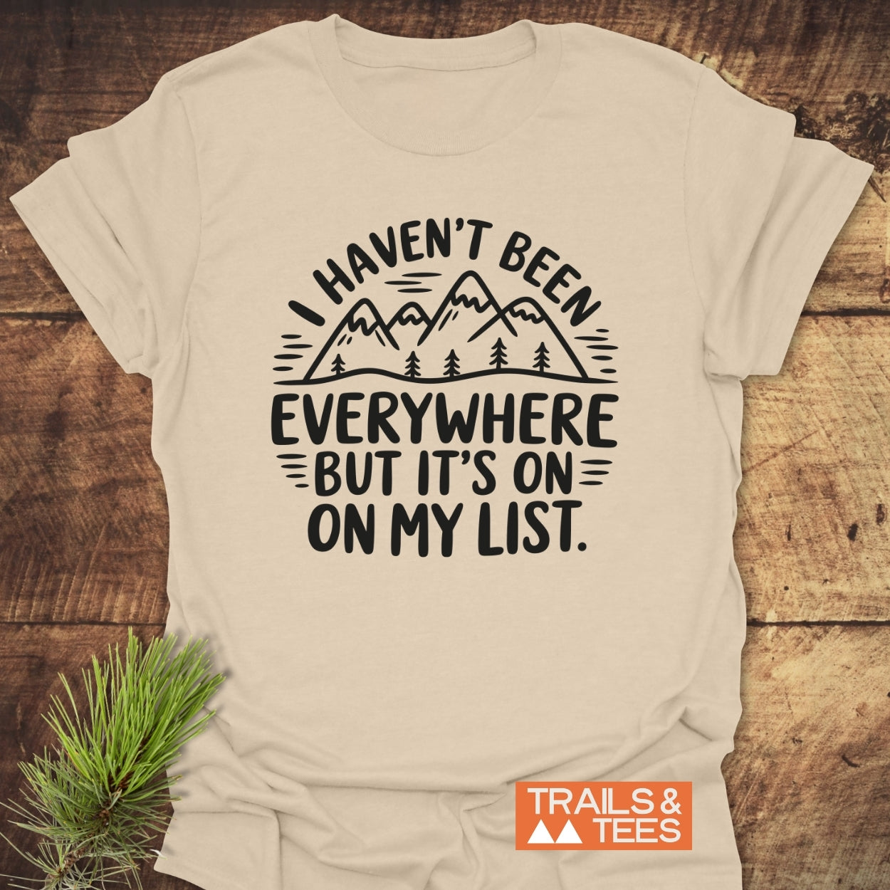 On My List T-Shirt featuring a mountain silhouette and trees graphic with travel-inspired text, laid flat with a pine branch. Perfect for outdoor enthusiasts, crafted for comfort and adventure.