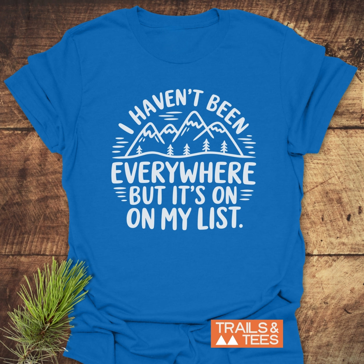 On My List T-Shirt featuring a white hand-drawn mountains and trees graphic with the slogan. Laid on wood, includes a small pine sprig and an orange Trails & Tees logo.
