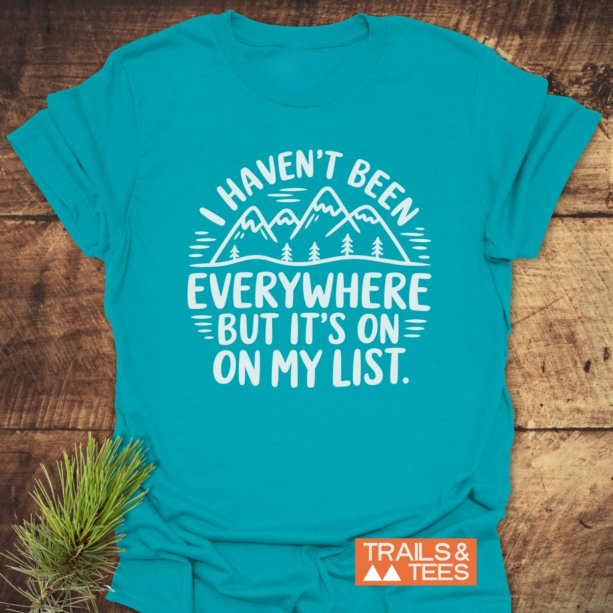On My List T-Shirt with a mountain and trees graphic, slogan “I haven’t been everywhere but it’s on my list,” and Trails & Tees logo, styled with a pine branch.