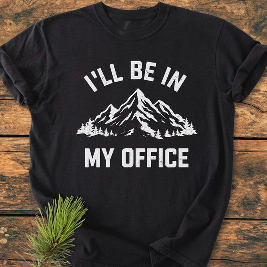 I'll Be In My Office - Vintage Wash Tee features a distressed white mountain and pine graphic on a black layout, displayed on a wooden table with a pine sprig accent.