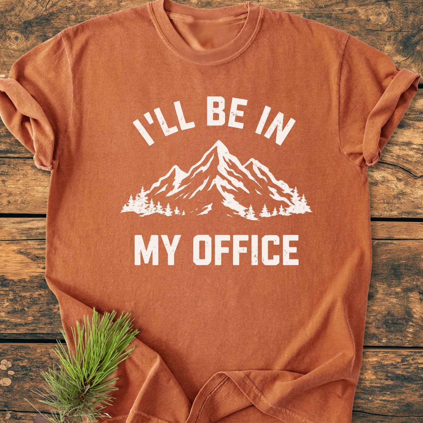 I'll Be In My Office - Vintage Wash Tee features a rust-orange design with a white mountain print, made from soft, durable garment-dyed cotton, laid flat on a wooden surface.