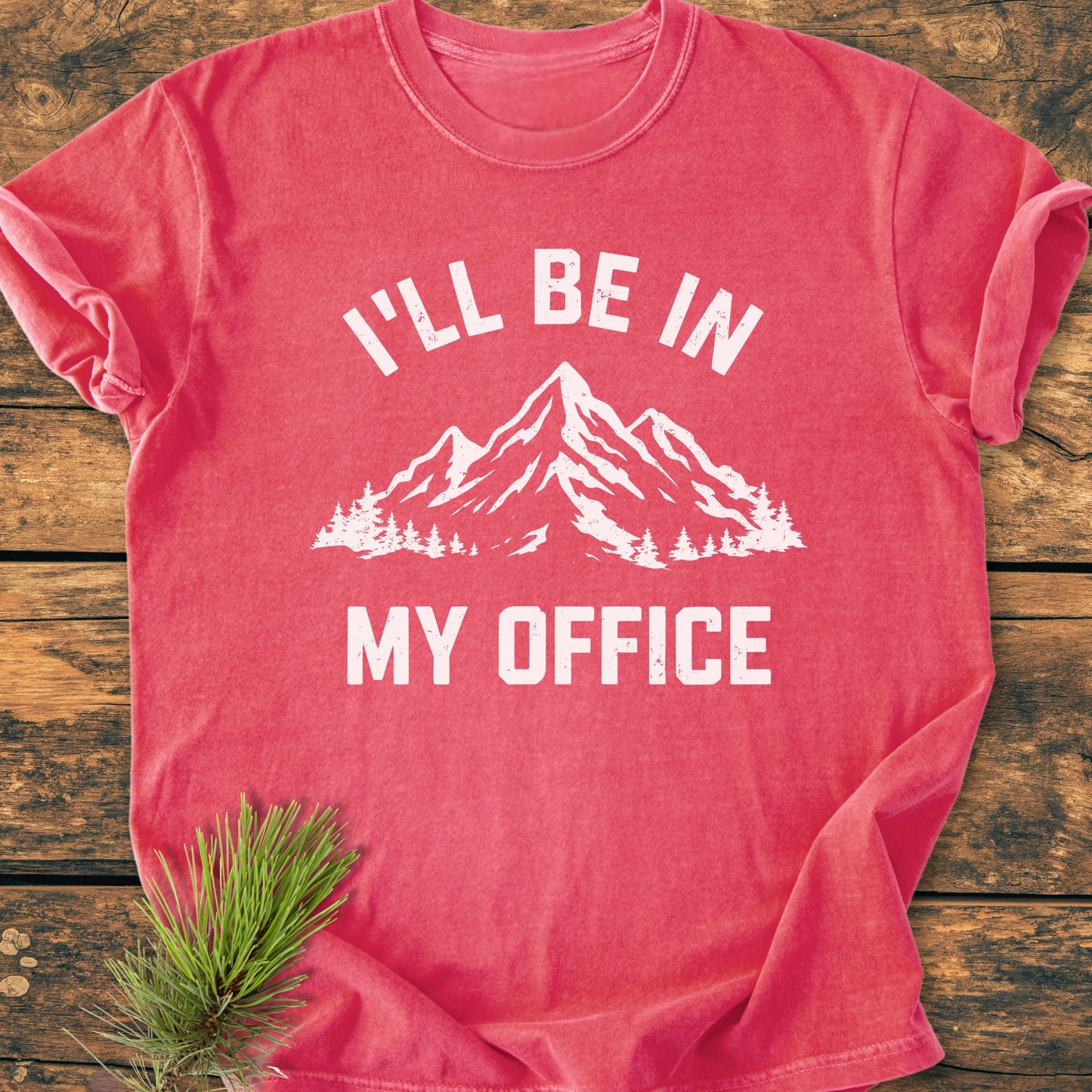 The I'll Be In My Office - Vintage Wash Tee features a mountain graphic and slogan on a faded-red shirt, laid on wood with a pine sprig under the hem for a rustic touch.